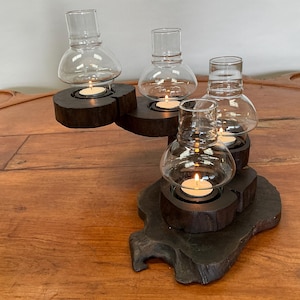 May include: A wooden candle holder with four glass candle holders. The candle holder is made of dark wood and has a natural, rustic look. The glass candle holders are clear and have a round shape. The candle holder is lit with four tea lights.