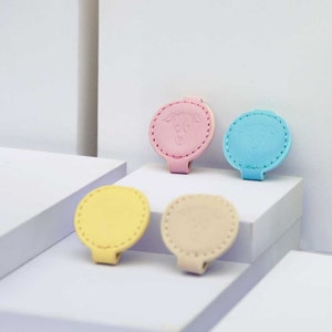 Pode incluir: Uma coleção de clipes coloridos com tema de cães. Os clipes são em tons pastel de amarelo, rosa, azul e bege. Cada clipe apresenta uma borda costurada e um design de rosto de cão. Os clipes estão dispostos em plataformas brancas.