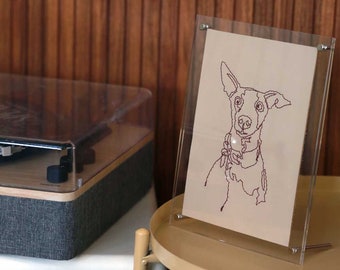 May include: A clear acrylic frame with a white mat holding a line drawing of a dog. The dog is brown and has a collar. The frame is sitting on a light brown table.