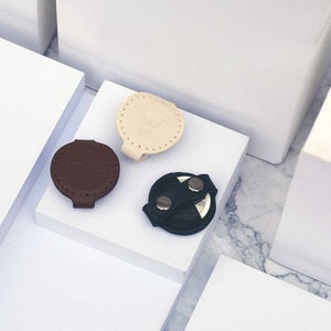 May include: Three round leather keychains in different colors: beige, brown, and black. The beige keychain has a stitched design and a small logo. The brown keychain has a stitched design. The black keychain has a strap with two silver snaps.