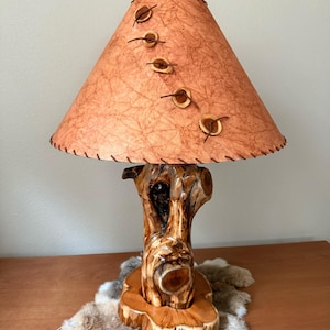May include: A rustic table lamp with a natural wood base and a textured, peach-colored lampshade. The shade is adorned with decorative wooden accents and stitched trim. Antlers are attached to the top of the lampshade. The lamp sits on a wooden table.