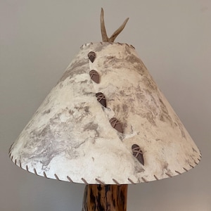 May include: A rustic table lamp with a conical lampshade. The shade is cream-colored with a marbled pattern and adorned with decorative brown accents. The base is made of natural wood, and the top features a pair of antlers.
