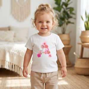 Könnte beinhalten: Ein weißes kurzärmeliges T-Shirt mit einem großen Buchstaben A als Grafik auf der Brust. Der Buchstabe besteht aus verschiedenen rosa, roten und weißen Blüten, darunter Rosen und Gänseblümchen, die in Form des Buchstabens A angeordnet sind.