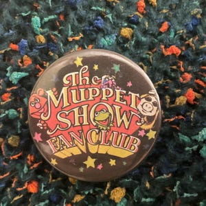 May include: A round pinback button featuring a black background with the text The Muppet Show Fan Club in red and yellow lettering. The design includes illustrations of Fozzie Bear, Kermit the Frog, and Miss Piggy, accented by small colorful stars.