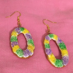 May include: A pair of gold dangle earrings with a donut-shaped charm. The donut is decorated with purple, green, and yellow icing.