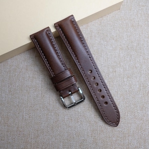Puede incluir: Correa de reloj de cuero marrón de dos piezas con hebilla plateada. La correa tiene un borde cosido y múltiples orificios para ajuste. El cuero parece liso y el color es un marrón oscuro intenso.