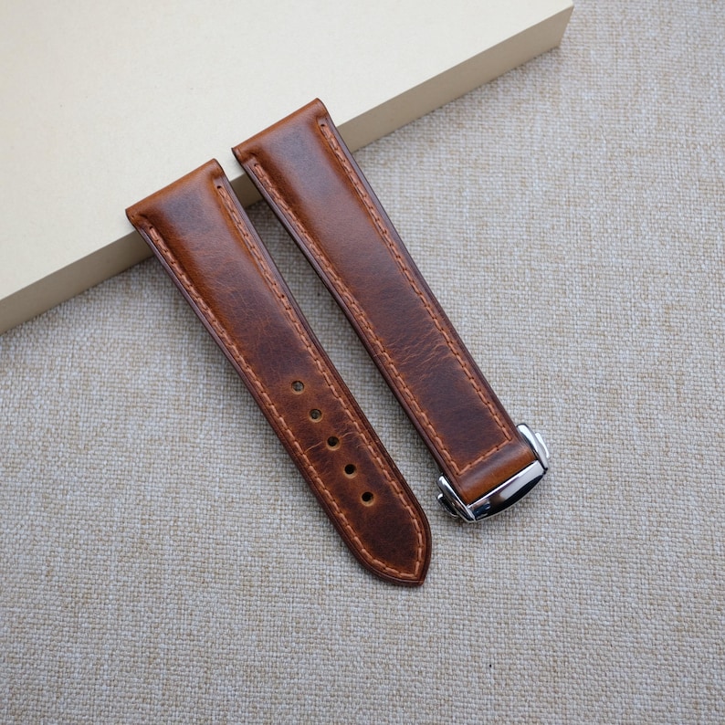 May include: Two brown leather watch straps with silver buckles. The straps are laid out flat on a light brown fabric surface.