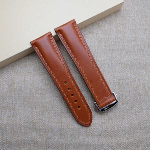 Puede incluir: Dos correas de reloj de cuero marrón con costuras color crema. Una correa tiene cinco agujeros y la otra una hebilla plateada. Las correas están hechas de cuero liso y están diseñadas para un reloj.