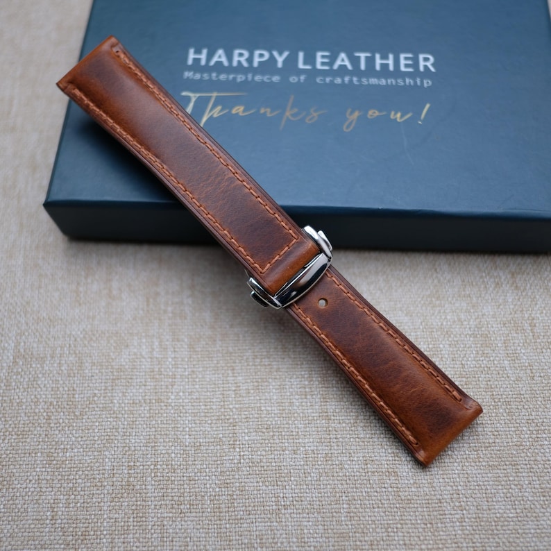 May include: A brown leather watch strap with a silver buckle. The strap has a stitched edge and is on a beige fabric background. The text "HARPY LEATHER Masterpiece of craftsmanship Thanks you!" is visible on a blue box in the background.