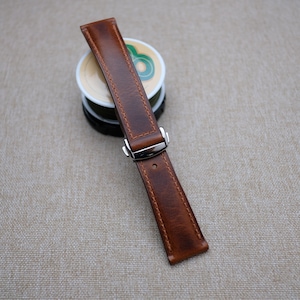 May include: A brown leather watch strap with a silver buckle. The strap has a stitched edge and a distressed look.