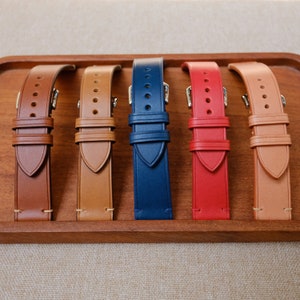 K&ouml;nnte beinhalten: F&uuml;nf Lederarmb&auml;nder in verschiedenen Farben: Braun, Beige, Dunkelblau, Rot und Beige. Die Armb&auml;nder werden auf einem Holztablett pr&auml;sentiert.