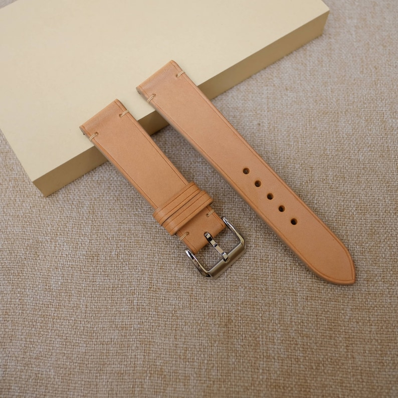 K&ouml;nnte beinhalten: Zwei hellbraune Lederarmb&auml;nder mit silbernen Schlie&szlig;en. Die Armb&auml;nder liegen auf einer beigefarbenen Stoffoberfl&auml;che.
