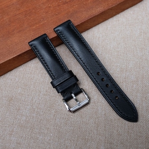 May include: Black leather watch strap with a silver buckle. The strap is stitched with a contrasting thread and has multiple holes for adjustment. The leather appears smooth and durable, suitable for various watch styles.