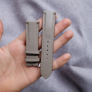 May include: Two grey leather watch straps with stitching. The strap on the left has a buckle and the strap on the right has a pointed end.