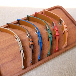 K&ouml;nnte beinhalten: Sechs Lederarmb&auml;nder in verschiedenen Farben, darunter Beige, Braun, Blau, Gr&uuml;n und Rot, auf einer Holzoberfl&auml;che pr&auml;sentiert.