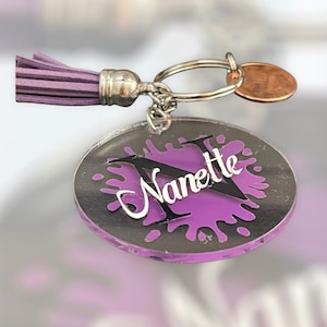 May include: Purple and black acrylic keychain with a splatter paint design and the name "Nanette" in white lettering.