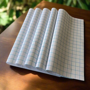 May include: A stack of white cross-stitch fabric sheets with a blue grid pattern. The sheets are fanned out on a wooden surface, with the grid lines clearly visible. The fabric is folded, creating a layered effect.