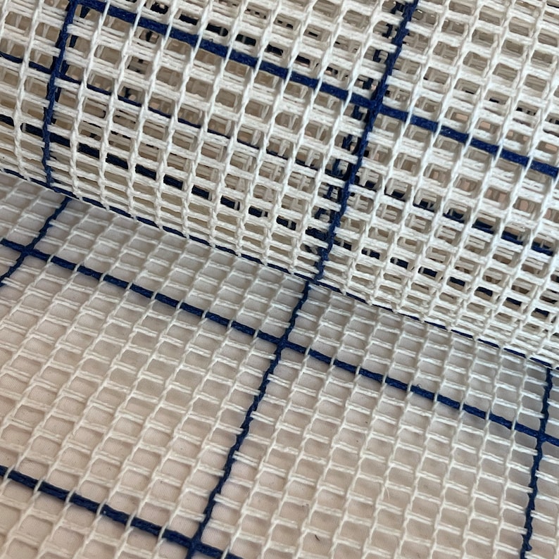 May include: A stiff, off-white rug hooking canvas featuring a grid pattern formed by dark navy blue lines. The fabric has a distinct open-weave structure with evenly spaced square holes, designed for needlepoint, latch hook, or embroidery projects.