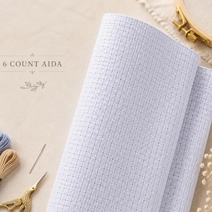 Puede incluir: Un rollo de tela Aida de color azul claro de 6 puntos con un patrón de cuadrícula distintivo para punto de cruz. La tela se muestra junto a tres madejas de hilo de bordar en blanco, azul y beige, una aguja y tijeras doradas de cigüeña.