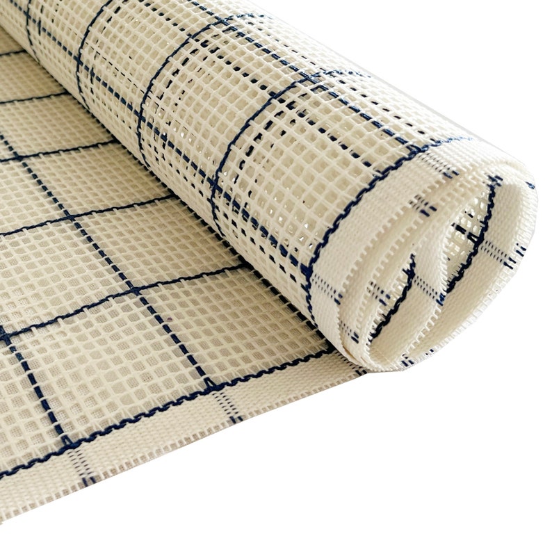 Op de afbeelding: Een rol off-white stramien voor handwerken met een ruitpatroon van dunne, donkerblauwe lijnen. De stof heeft een stijve, open geweven structuur met gelijkmatig verdeelde gaten, ontworpen voor naaldwerk, knoopwerk of het maken van tapijten.