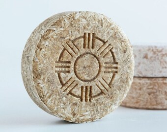 May include: Three round, solid, beige shampoo bars. The bar in the front has a circular design with a sun-like symbol in the center.