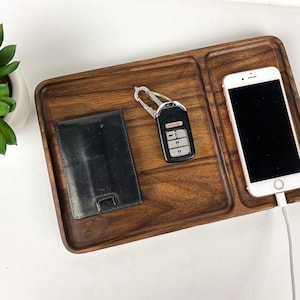 May include: A wooden tray with two compartments, one for a phone and one for keys and a wallet. The phone is charging with a white cable. The tray is made of dark wood.