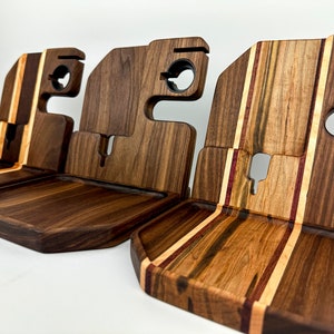 May include: Three wooden phone stands with different wood grain patterns. The stands have a raised edge to hold a phone and a cutout for charging cables. The stands are made of walnut and maple wood.