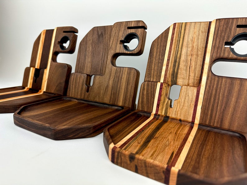 May include: Three wooden phone stands with different wood grain patterns. The stands have a raised edge to hold a phone and a cutout for charging cables. The stands are made of walnut and maple wood.