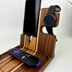 May include: A wooden phone and watch stand with a tray for a remote control. The stand is made of walnut wood with a striped pattern. The phone stand is on the left, the watch stand is on the right, and the tray is in the center. A black phone, a black watch, and a black remote control are on the stand.