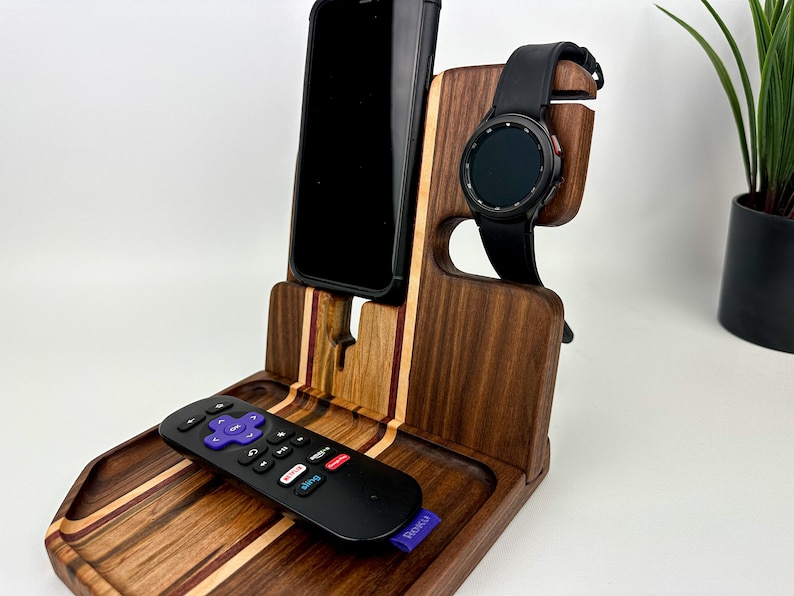 May include: A wooden phone and watch stand with a tray for a remote control. The stand is made of walnut wood with a striped pattern. The phone stand is on the left, the watch stand is on the right, and the tray is in the center. A black phone, a black watch, and a black remote control are on the stand.