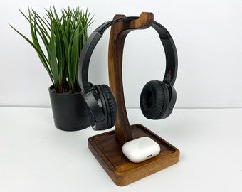 May include: A wooden headphone stand with a curved design, holding black headphones. A white charging case rests on the base. A potted green plant is in the background. The stand is made of dark wood.