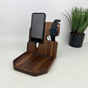 May include: A wooden phone and watch stand with a small tray. The stand is made of dark walnut wood and has a slot for a phone and a watch. The tray is perfect for holding keys, jewelry, or other small items.