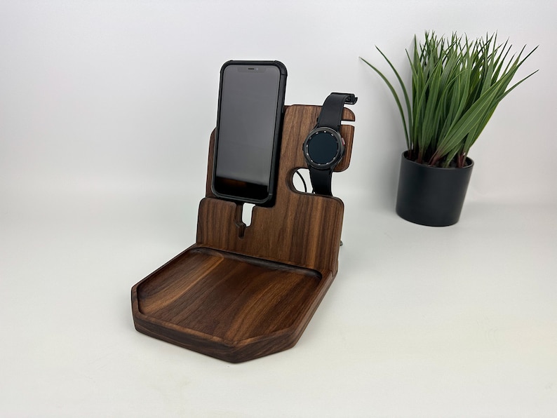 May include: A wooden phone and watch stand with a small tray. The stand is made of dark walnut wood and has a slot for a phone and a watch. The tray is perfect for holding keys, jewelry, or other small items.