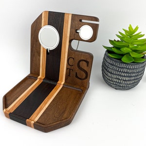 May include: A wooden charging station with a walnut and cherry wood finish. It has two charging pads for a watch and phone, a small tray, and the initials "CST" engraved on the side.