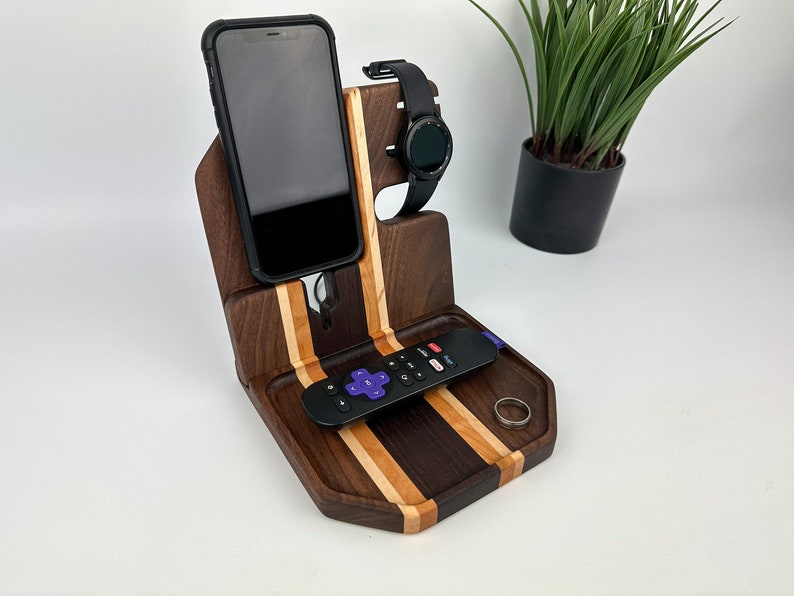 May include: A wooden phone and watch stand with a tray for a remote control and a ring. The stand is made of walnut and maple wood and has a striped pattern.