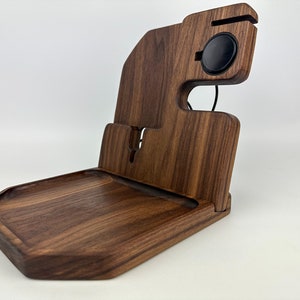 May include: A wooden charging station with a tray, a phone stand, and a wireless charging pad for a smartwatch. The station is made of dark brown walnut wood.