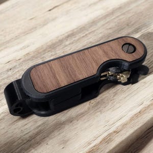 Peut inclure: Un organiseur de clés noir avec un incrustation en bois de noyer. Le porte-clés a un design élégant et moderne avec une vis en métal et peut contenir plusieurs clés. Le produit est présenté sur une surface en bois clair.