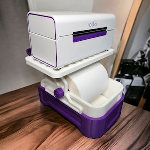 May include: A white and purple label printer with a roll of white label paper. The printer has the word "rollo" printed on the front.