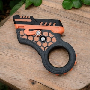 May include: A black and orange 3D-printed device with a ring handle. The device has a geometric design with a honeycomb pattern and a trigger-like mechanism. The object is resting on a wooden surface.