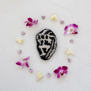 May include: A black and white embroidered design of a person's face surrounded by pink and white orchid flowers on a white background.