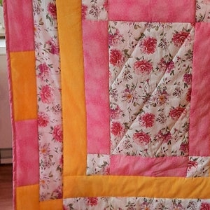 Könnte beinhalten: Eine rosafarbene und gelbe Patchwork-Decke mit einem Blumenmuster. Die Decke besteht aus Quadraten und Rechtecken in verschiedenen Größen und Farben.