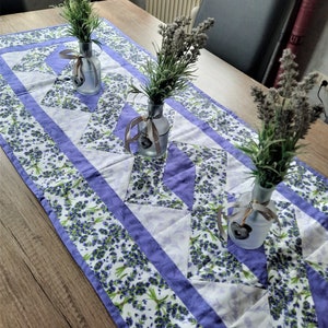 Op de afbeelding: Een paarse en witte tafelrunner met een lavendelbloemmotief. Drie glazen vazen met lavendeltakken zijn op de runner gerangschikt.