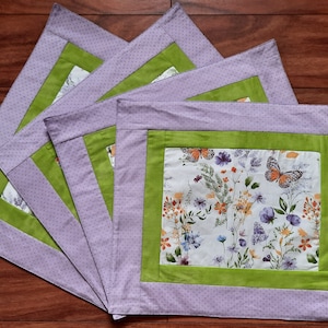 Könnte beinhalten: Set mit vier lila und grünen Platzsets mit einem Blumen- und Schmetterlingsmuster. Die Platzsets haben einen weißen Hintergrund mit grünen Rändern und einem Blumenmuster mit Schmetterlingen.