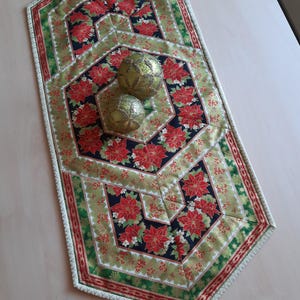 Könnte beinhalten: Ein gesteppter Weihnachtstischläufer mit einem sich wiederholenden Muster aus roten Weihnachtssternen auf schwarzem Grund, umrahmt von grünen und roten Bordüren. Zwei goldene Dekokugeln liegen darauf.