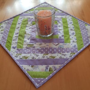 Könnte beinhalten: Ein quadratischer, gesteppter Tischset mit einer Kerze in einem Glas. Das Tischset hat ein Patchwork-Design mit Stoffen in Lavendel, Grün und Weiß. Die Kerze ist pfirsichfarben. Das Tischset liegt auf einer Holzoberfläche.