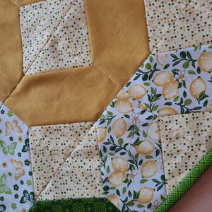 May include: A quilted table runner featuring a patchwork design with solid mustard yellow fabric, a cream fabric with small green dots, a white fabric with yellow lemons and green leaves, and a white fabric with green and yellow butterflies.