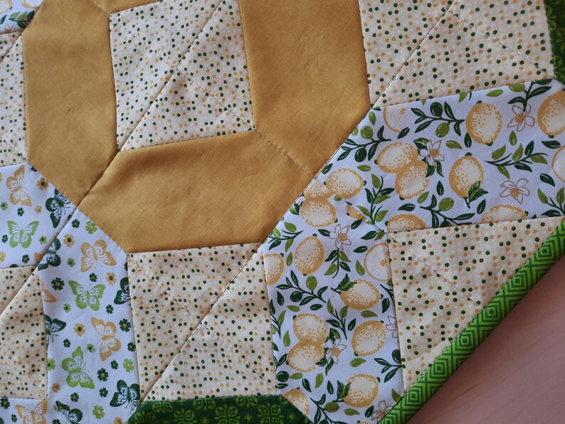 May include: A quilted table runner featuring a patchwork design with solid mustard yellow fabric, a cream fabric with small green dots, a white fabric with yellow lemons and green leaves, and a white fabric with green and yellow butterflies.
