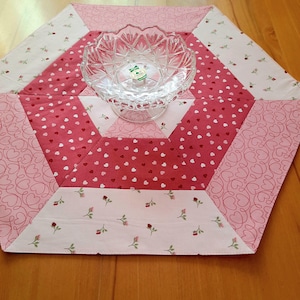 Puede incluir: Un cuenco de vidrio transparente con base de pedestal se encuentra sobre un camino de mesa hexagonal acolchado. El camino de mesa presenta un diseño de patchwork con telas rosas y blancas. Algunas secciones tienen un patrón de corazón, otras pequeñas rosas rojas.