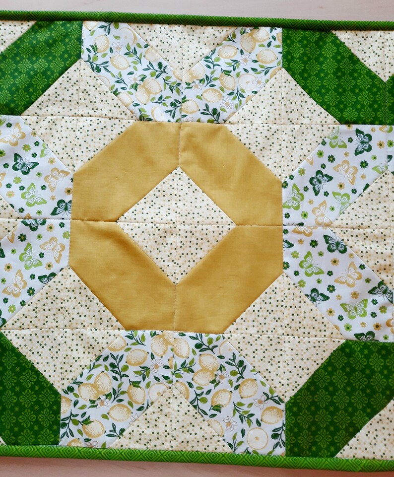 May include: A quilted fabric runner featuring a central mustard yellow octagon surrounded by cream fabric with small green dots. The design includes sections of white fabric printed with green and yellow butterflies and lemon patterns, bordered by green fabric.