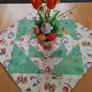 Könnte beinhalten: Ein quadratischer Tischläufer zum Thema Ostern mit einem Patchwork-Design. Der Läufer hat ein hellgrünes und cremefarbenes Blumenmuster mit Bildern von Hasen, Eiern und Blumen. Ein kleiner dekorativer Korb mit Eiern und einer Hühnerfigur befindet sich in der Mitte, mit orangen Tulpen.